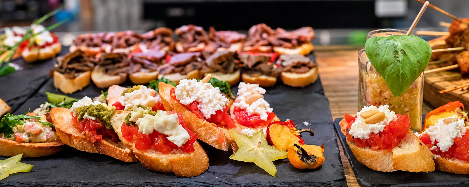 Des tranches de pain garnies de tomates, de fromage frais et de pesto sont présentées sur des ardoises, à côté d'un verre décoré d'une feuille de basilic.
