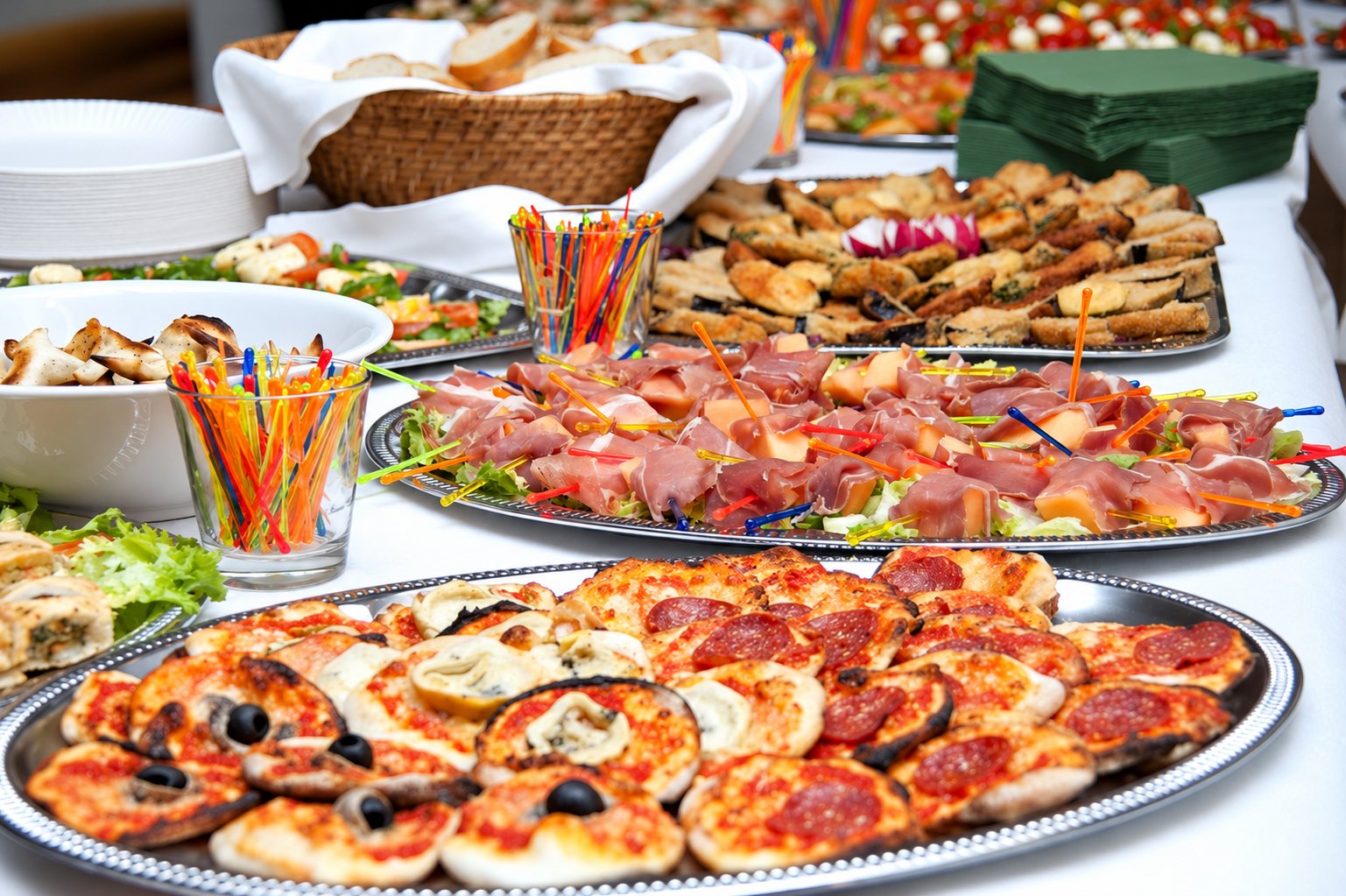 Table de buffet couverte d'une nappe blanche avec des plateaux de mini pizzas, de jambon cru au melon et de canapés, accompagnés de piques colorés.