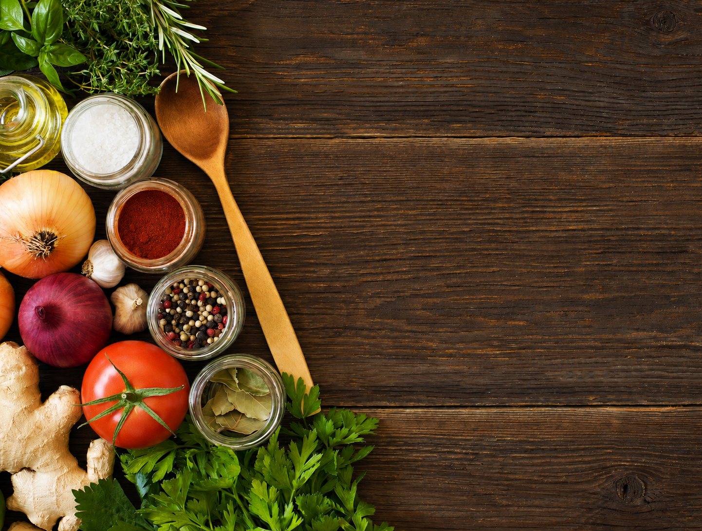 Des bocaux d'épices, une cuillère en bois, des oignons, de l'ail, une tomate, du gingembre et des herbes fraîches sont disposés sur une table en bois sombre.