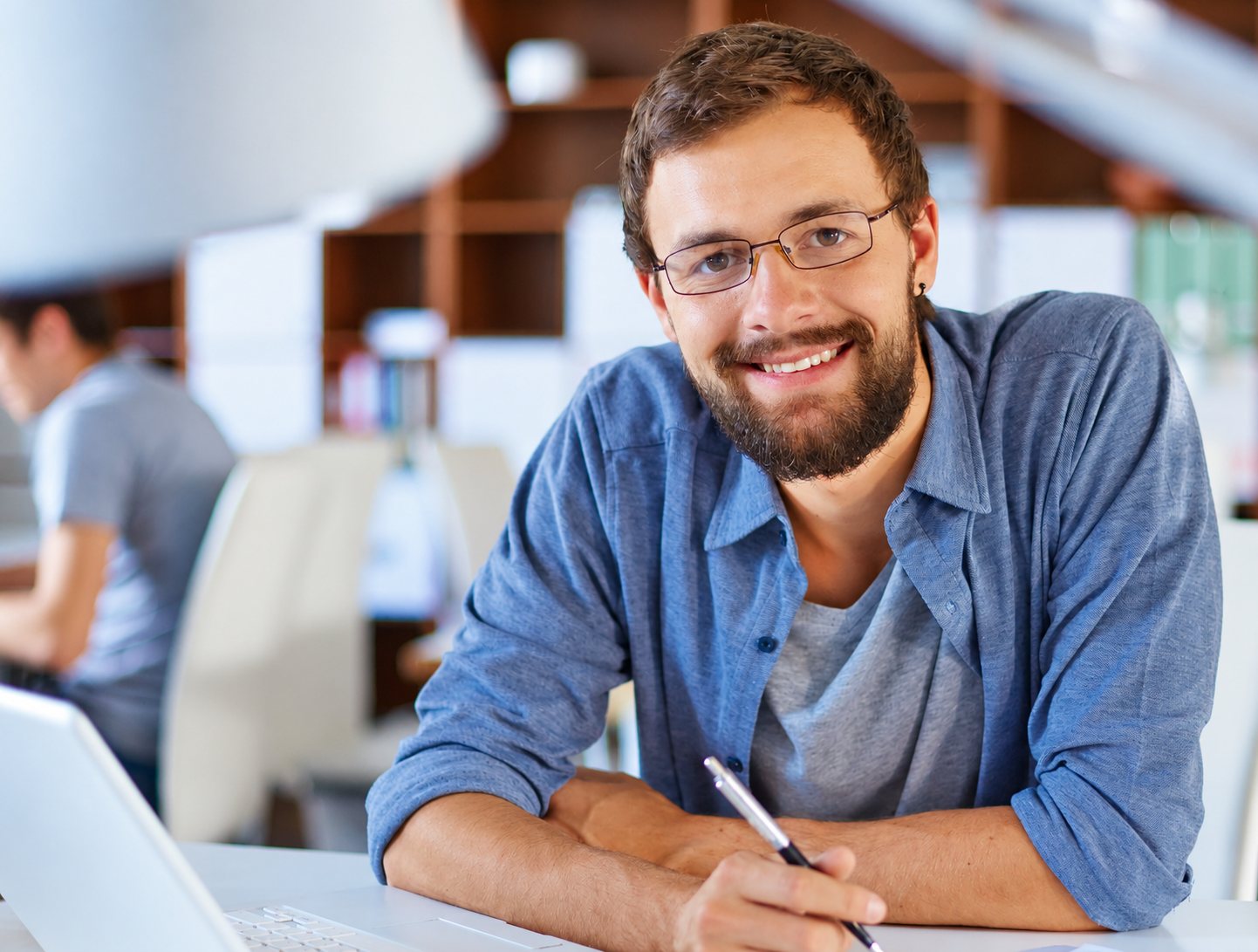 Un jeune homme barbu à lunettes sourit en tenant un stylo devant un ordinateur portable, dans un bureau lumineux où un collègue est flou à l'arrière-plan.