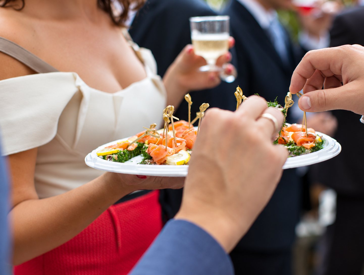 Lors d'une réception, des invités prennent des canapés au saumon fumé, citron et aneth sur une assiette blanche, un verre de vin blanc en arrière-plan.