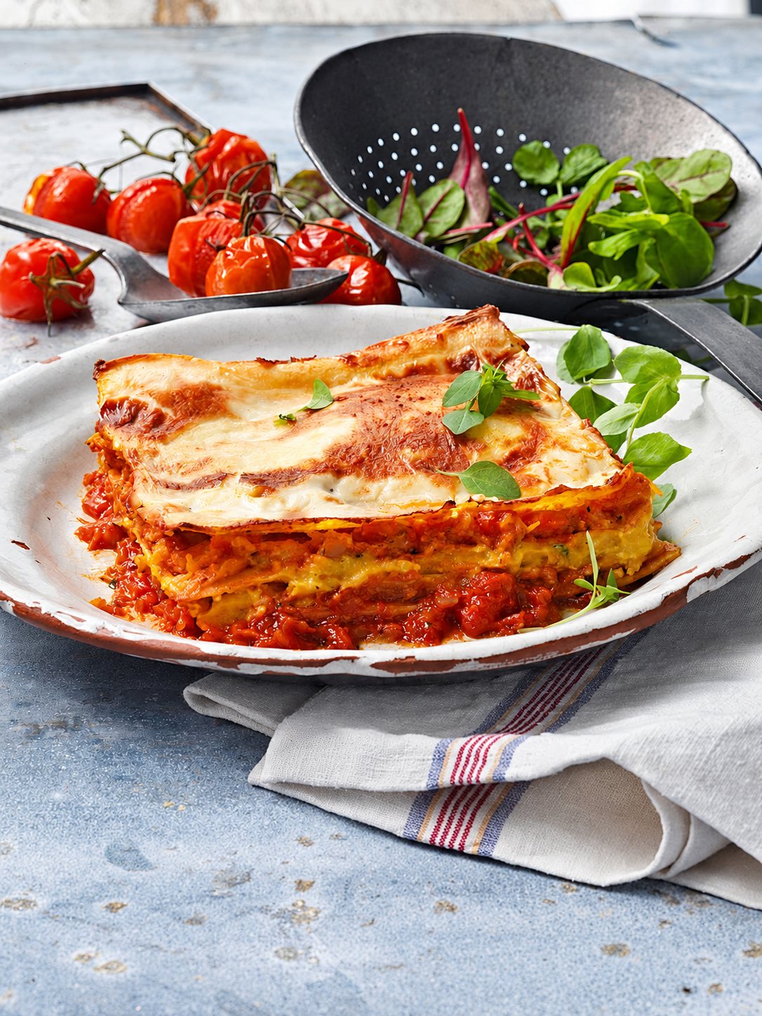 Une part de lasagnes gratinées dans une assiette émaillée, entourée de tomates cerises rôties, d'une passoire de jeunes pousses et d'un torchon rayé.